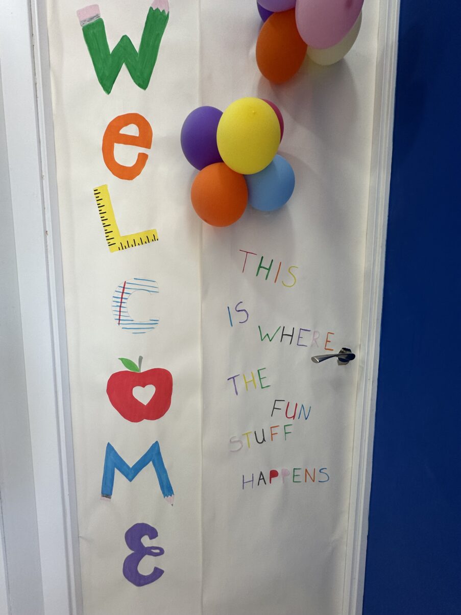 Puerta decorada con la palabra “Welcome”, globos de colores y el mensaje “This is where the fun stuff happens”