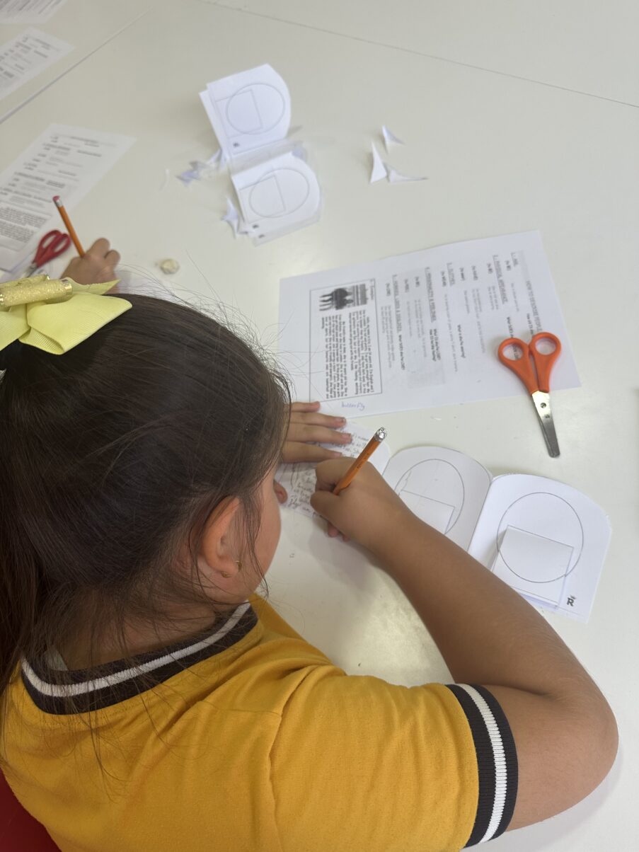 Alumna de la academia Royal English Valdemoro vista desde atrás mientras escribe en su librito de descripciones en inglés, utilizando una hoja de vocabulario como referencia.