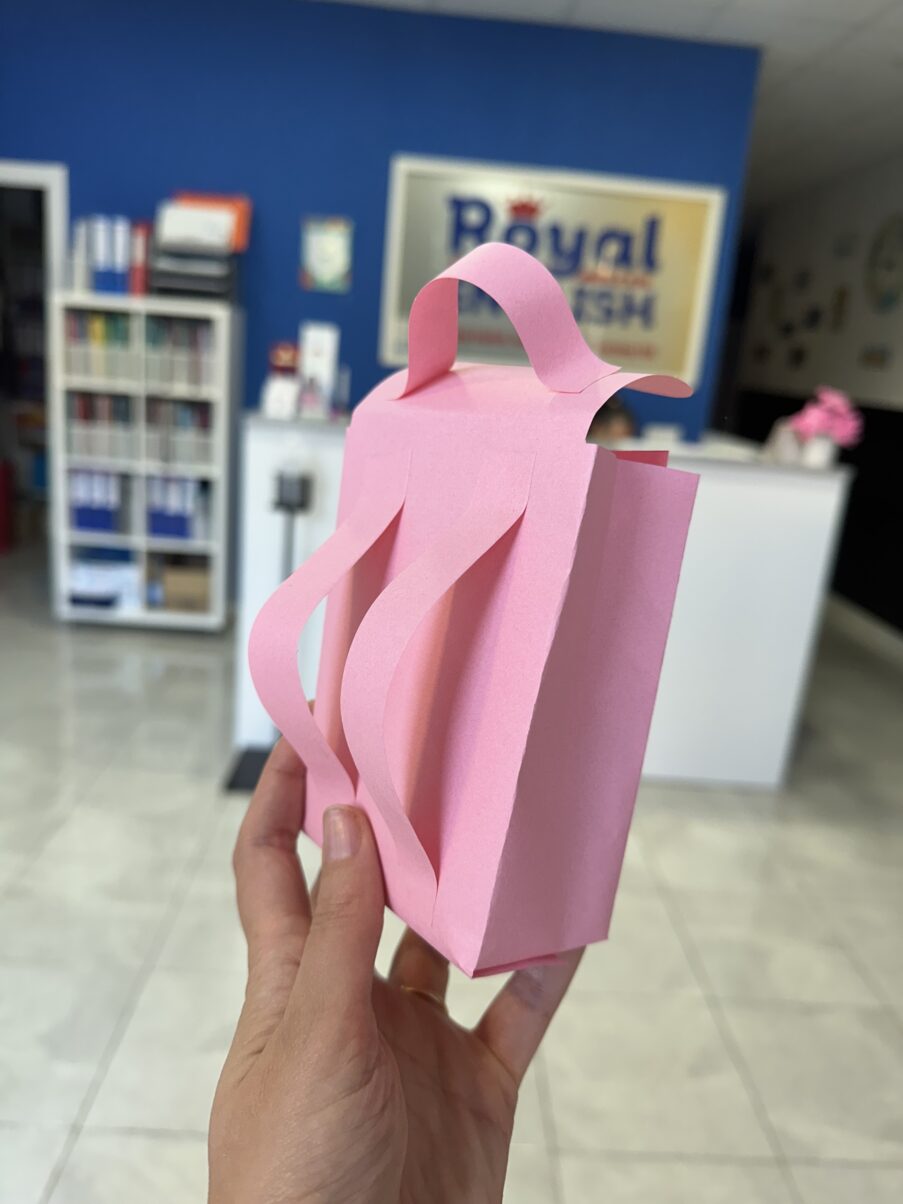 Mano sosteniendo una pequeña mochila escolar rosa hecha de papel, con correas pegadas. Al fondo se distingue de manera borrosa la recepción y el logotipo de Royal English.
