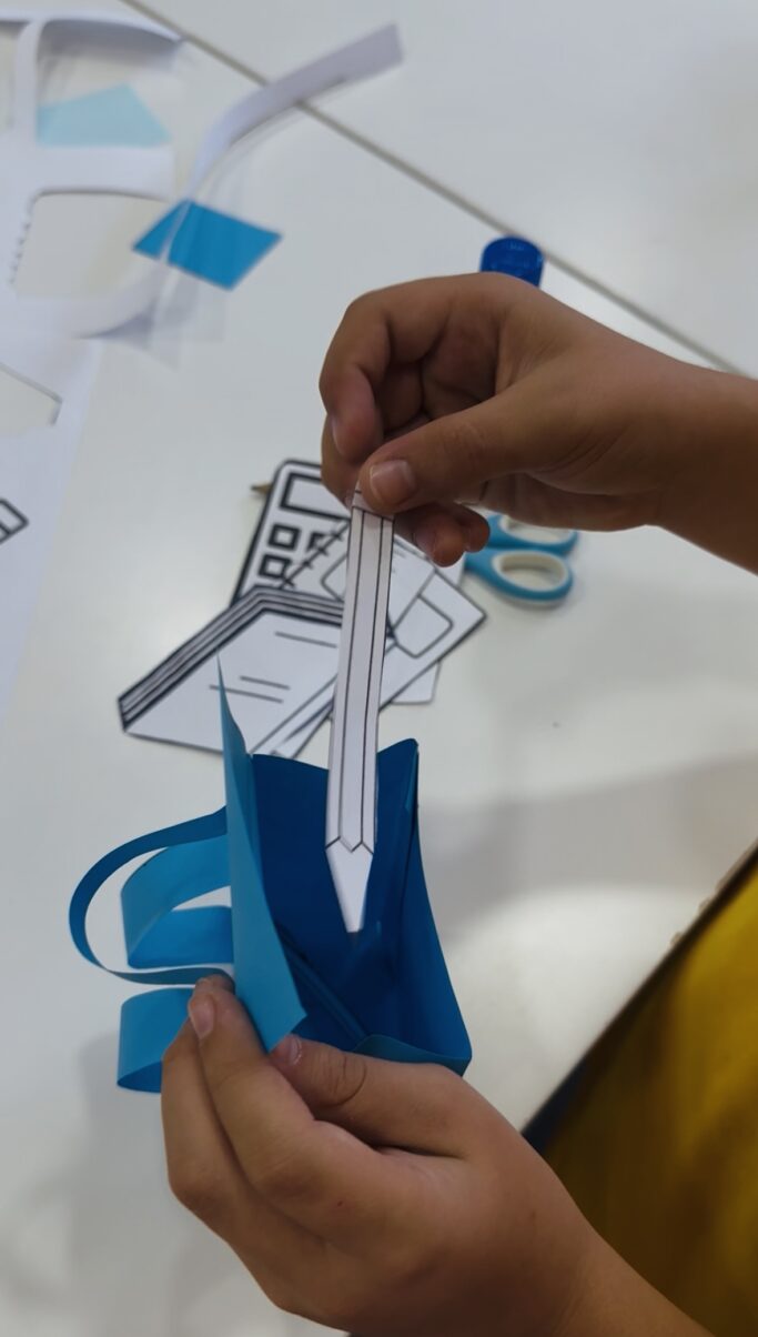 Estudiante sacando un lápiz de una pequeña mochila azul hecha de papel, con otros objetos escolares como un cuaderno, un libro, una calculadora, tijeras y pegamento visibles en el fondo.